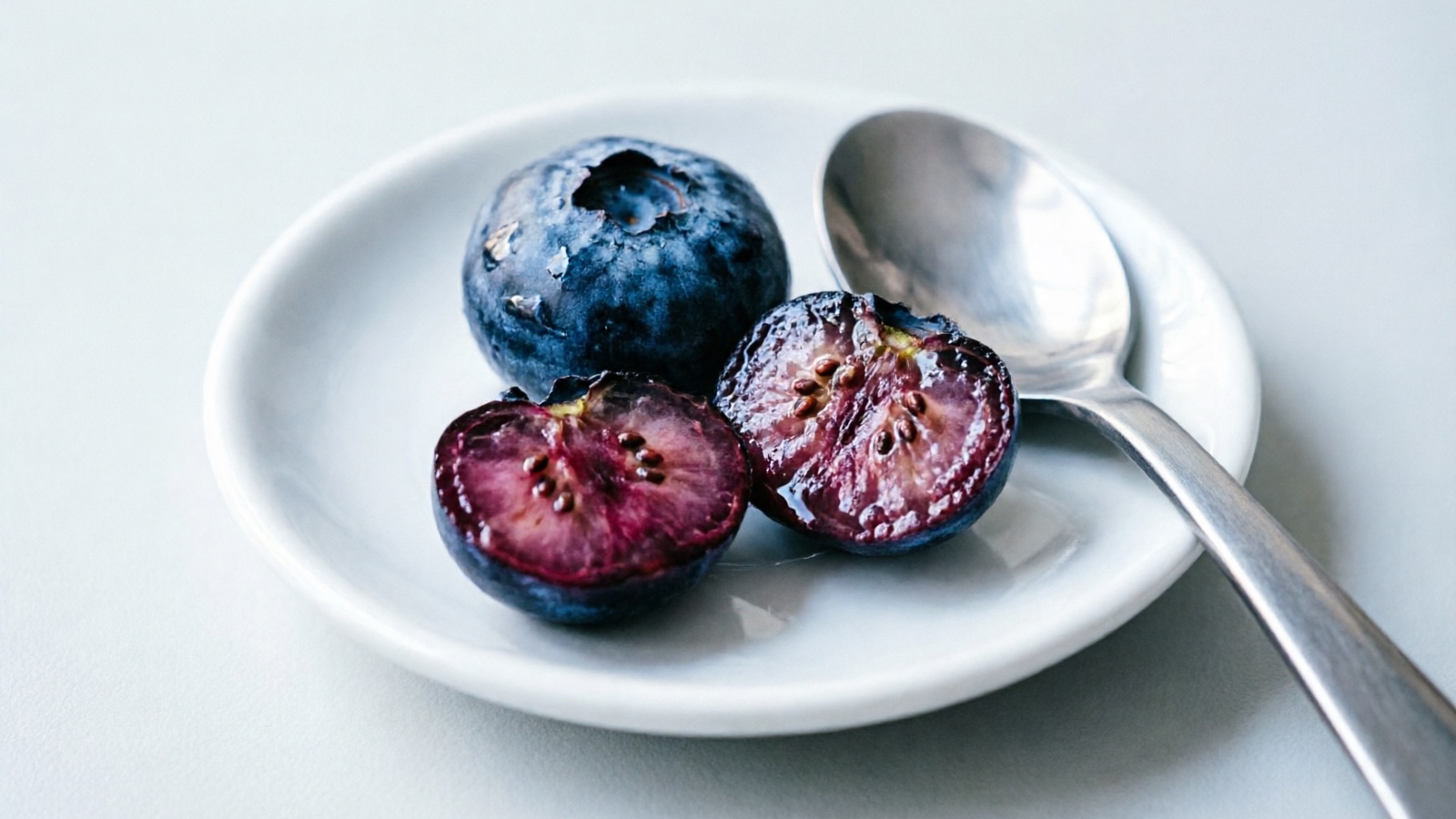 Two blueberries on a small dish, one whole and one halved, beside a teaspoon for size reference