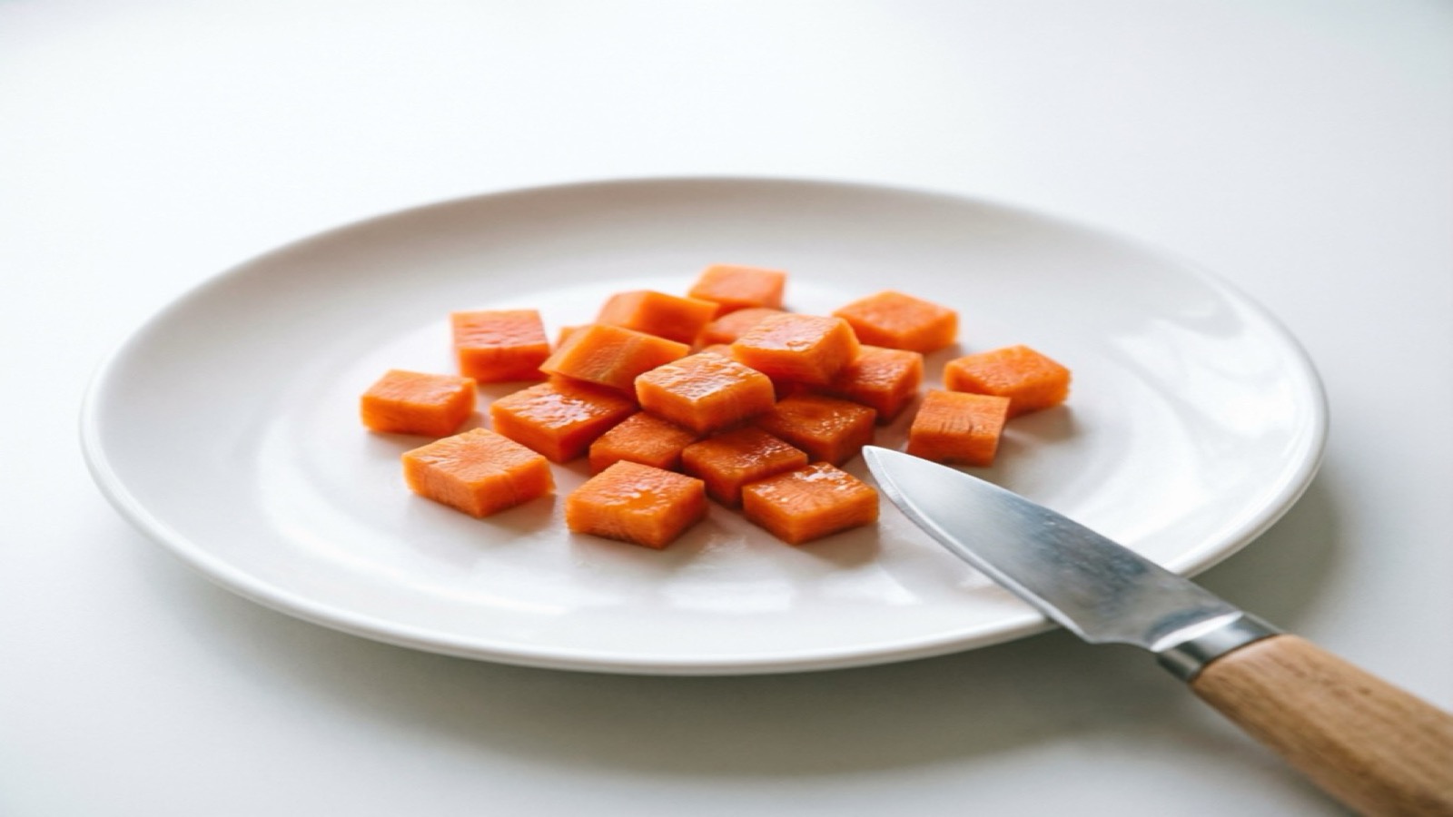 Close-up of carefully sliced thin carrot rounds and small bite-sized carrot pieces arranged on a clean wooden surface, with a small kitchen knife nearby